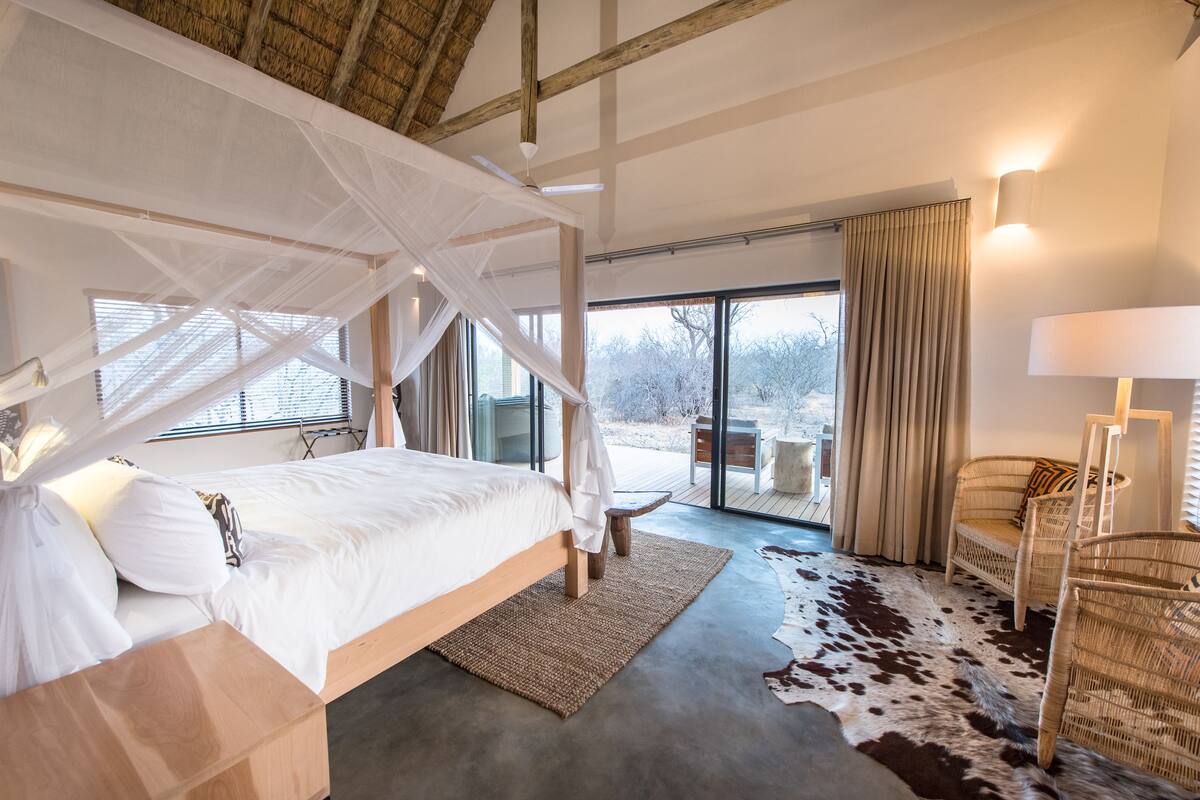 A bedroom with a canopy bed, a large window, a view of the outdoors, a wicker chair, and a cowhide rug.