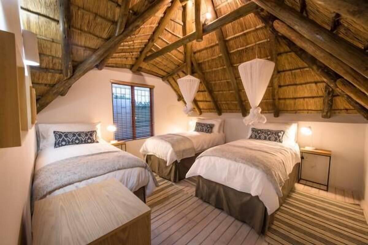 A cozy bedroom with three beds, a thatched roof, and a window with blinds.