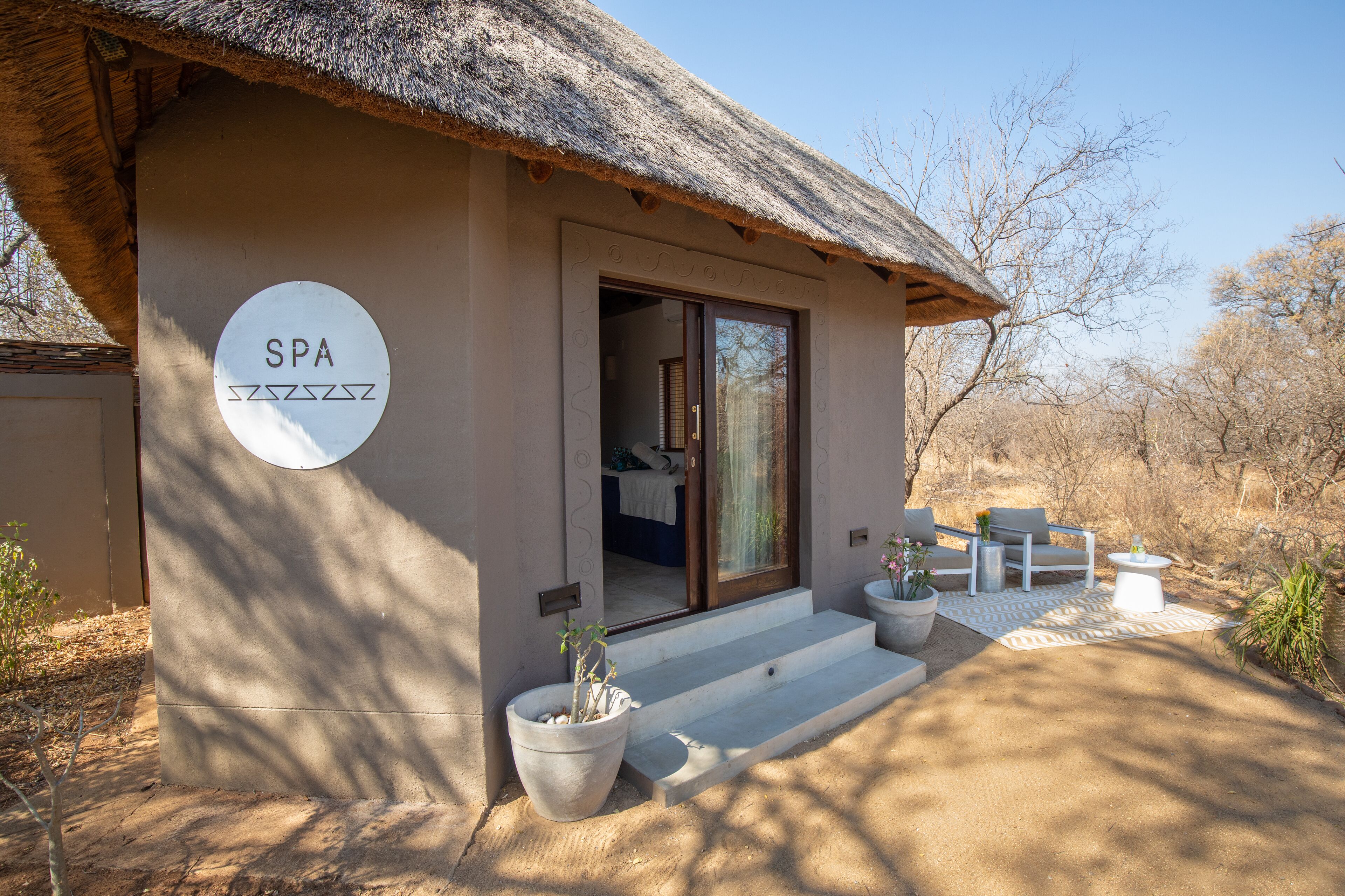 Un bâtiment abritant un spa, doté d’un toit de chaume, d’une enseigne indiquant « SPA » et d’une terrasse avec des meubles d’extérieur.