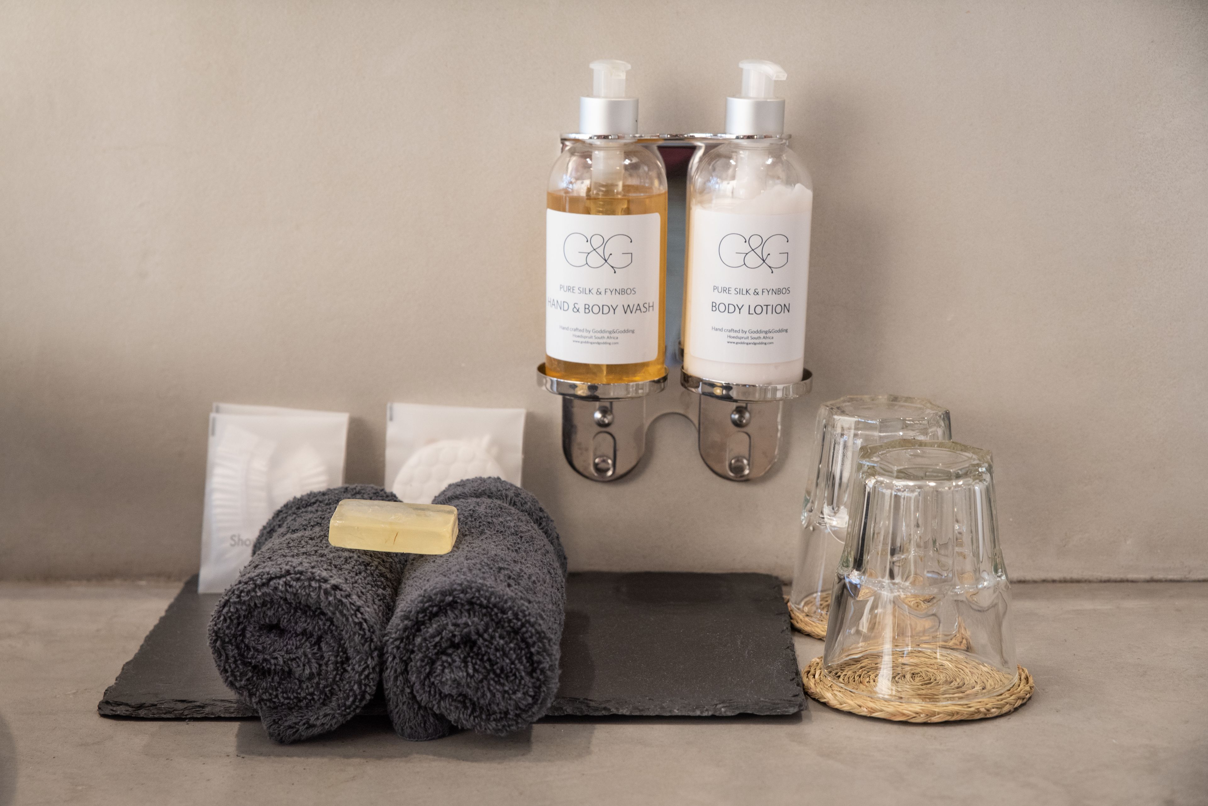 Two bottles of C&G Pure Silk & Fynbos hand and body wash, a pair of rolled towels, and three clear glasses on a tray.