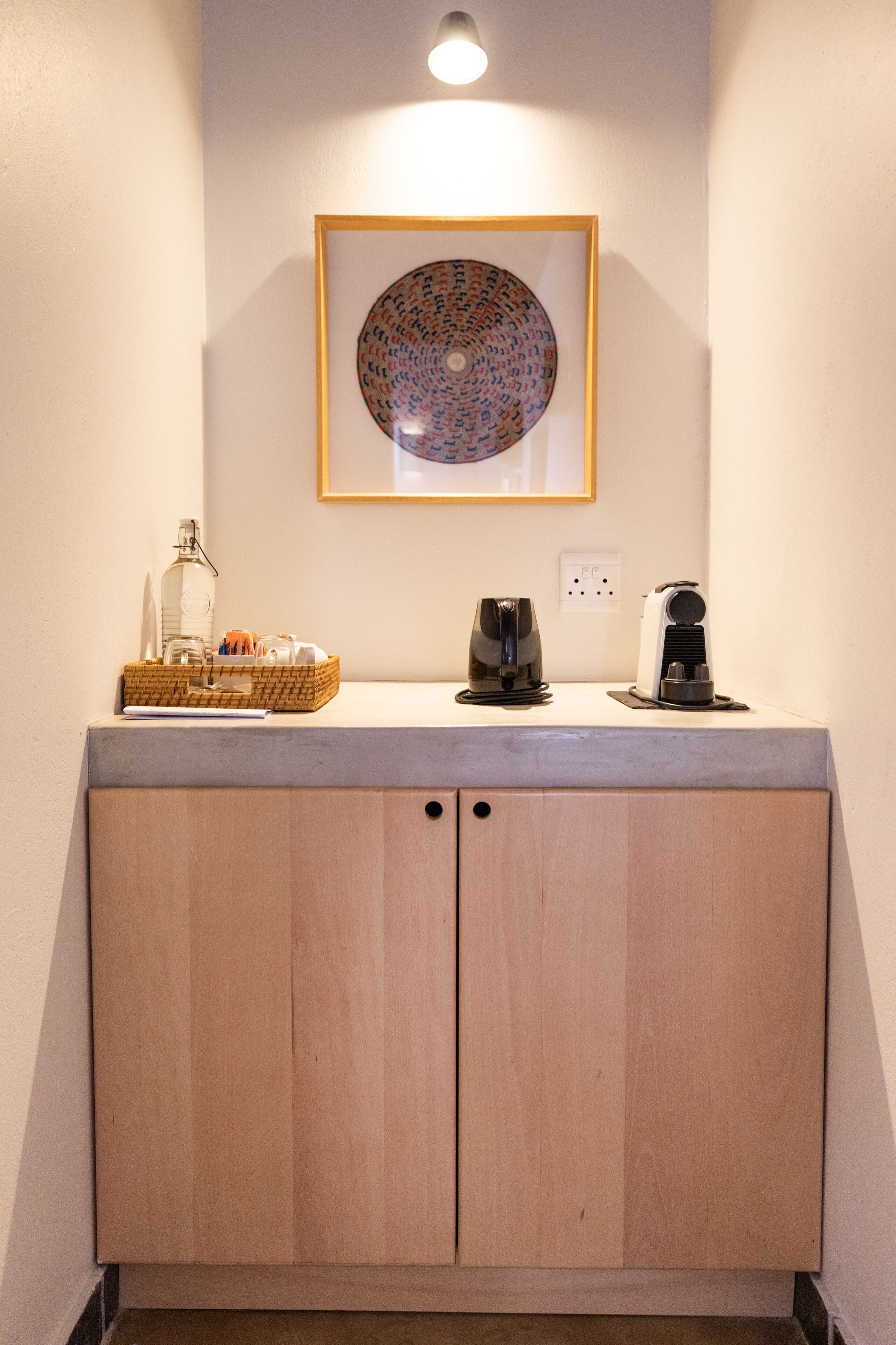 A kitchenette with a wooden cabinet, a framed artwork, a coffee maker, and a bottle on the countertop.