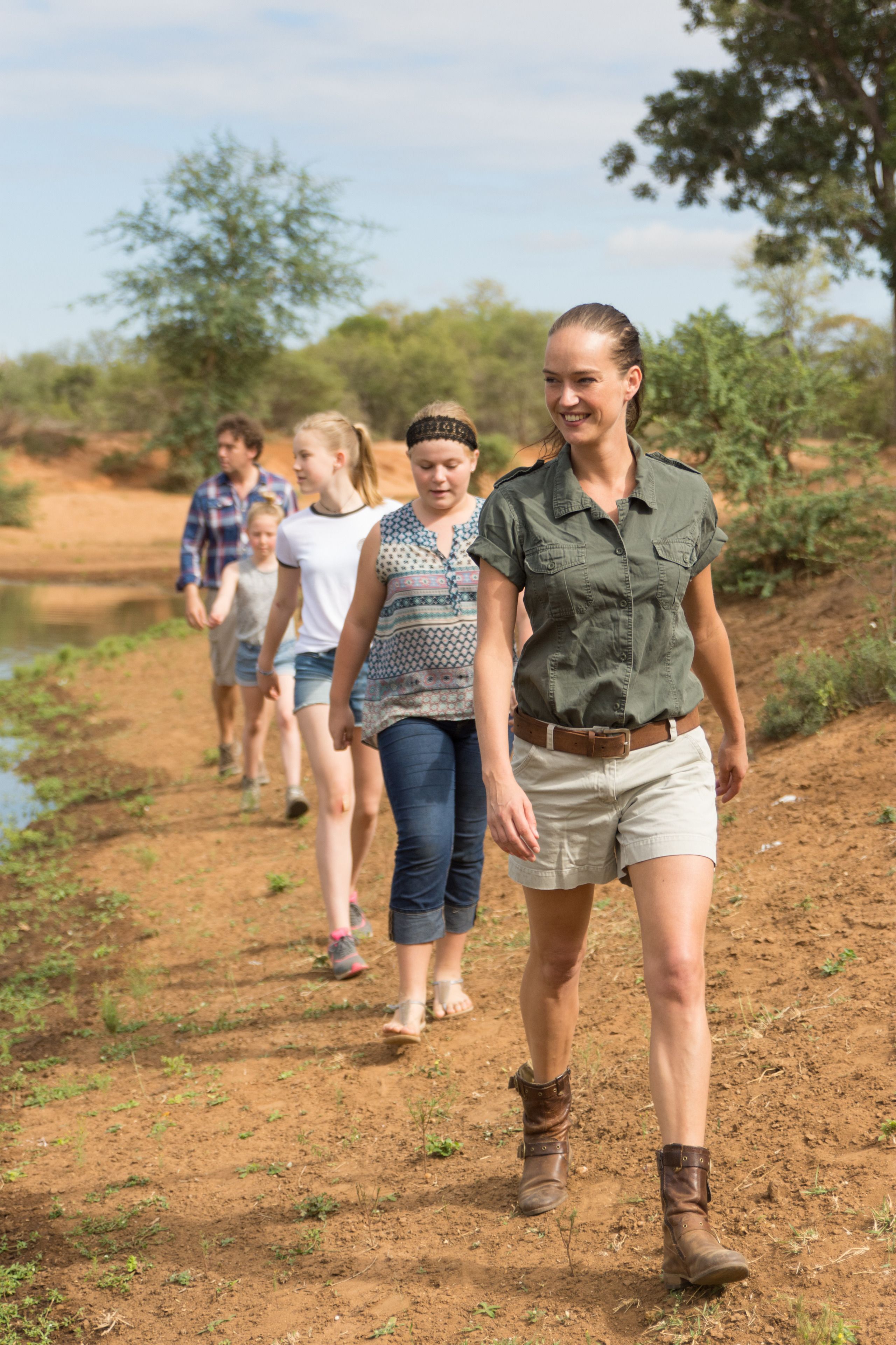 A group of people walking on a dirt path beside a body of water, surrounded by greenery.