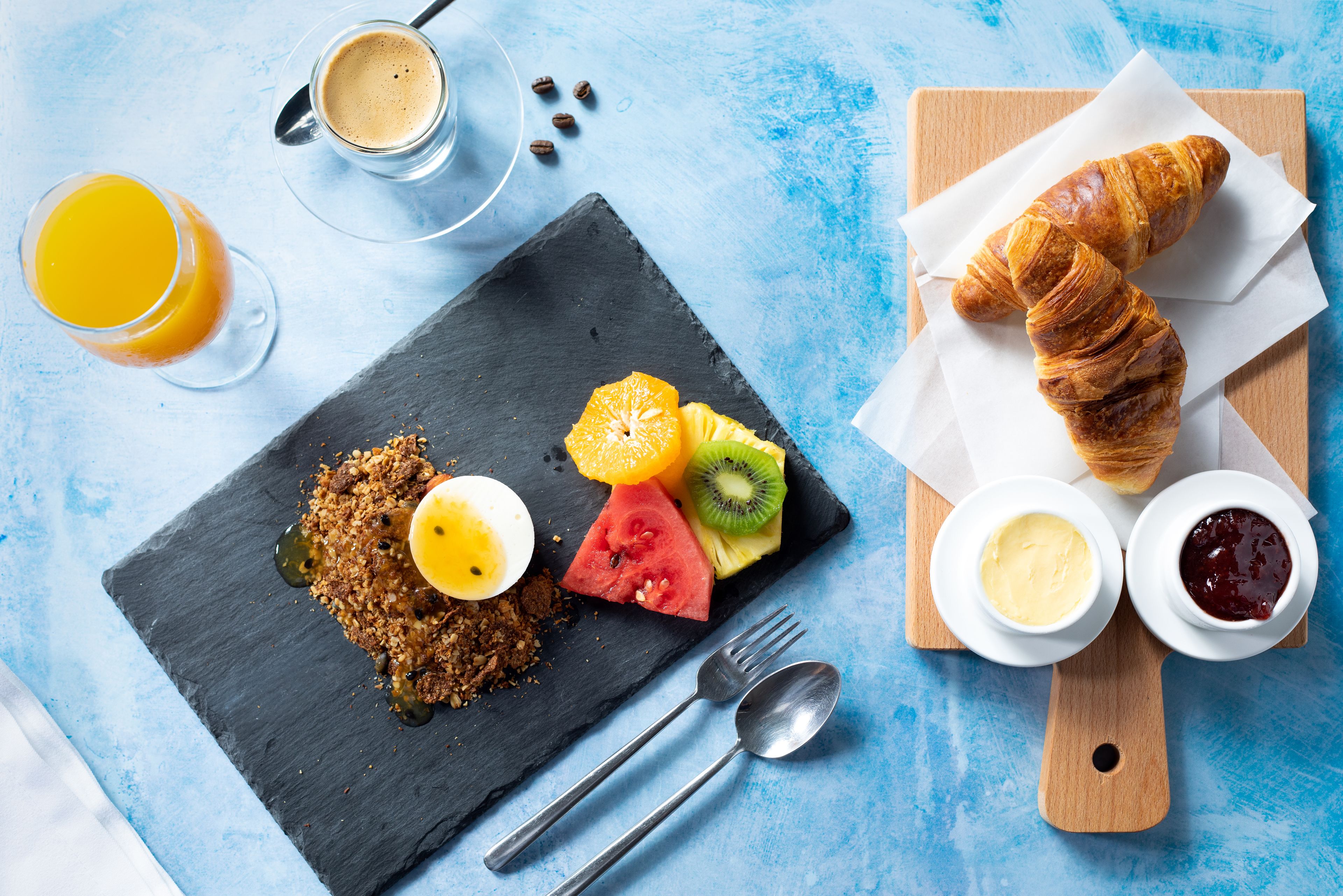A breakfast setup with croissants, fruit, and spreads on a slate board, along with a glass of orange juice and a cup of coffee.