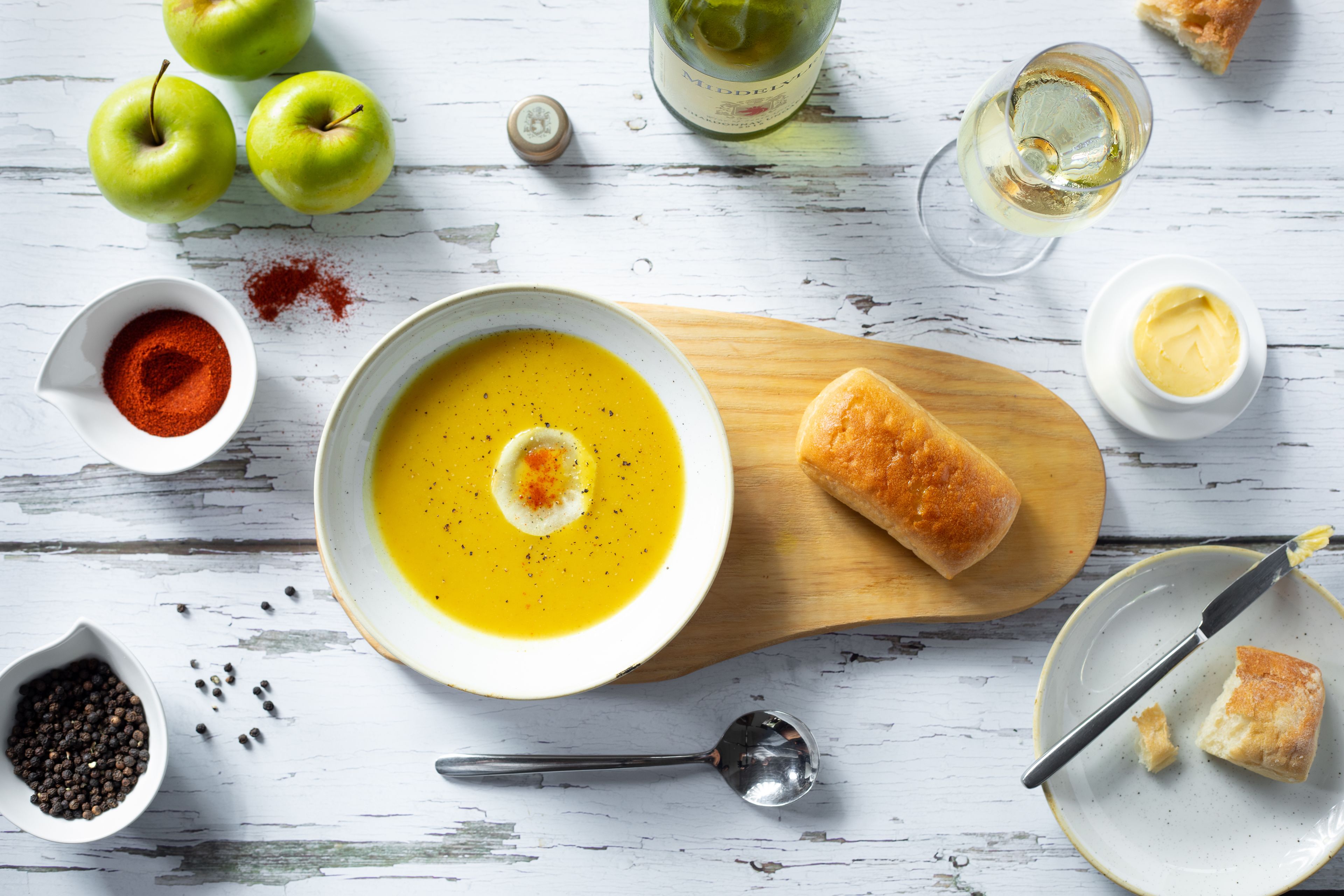 A bowl of soup with a slice of bread on a wooden board, surrounded by apples, a bottle of wine, and condiments.