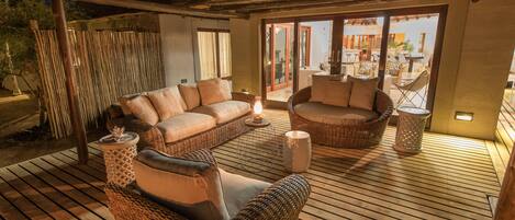 Una terraza de madera con muebles de mimbre, incluyendo sofás y una silla, y un vidrio que da al exterior.