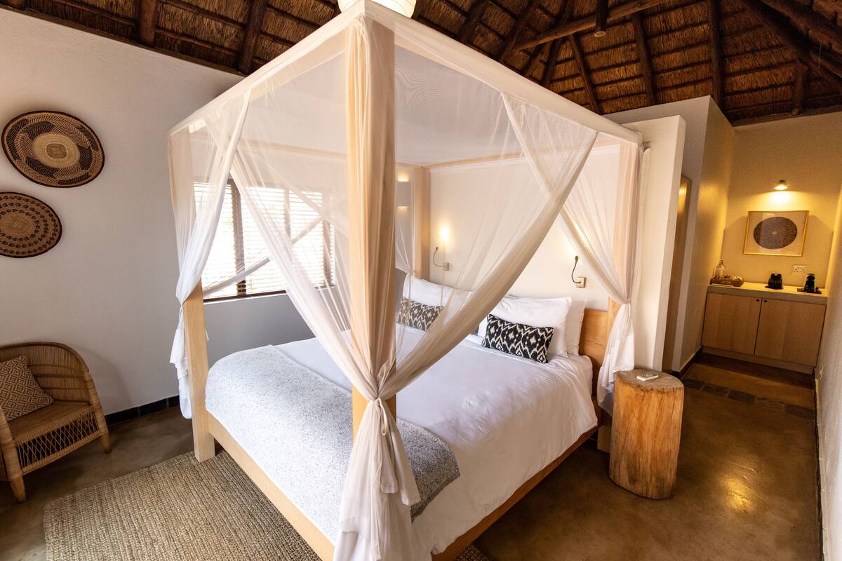 A four-poster bed with a canopy, a wicker chair, a wooden stool, and a kitchen area with cabinets and a countertop.