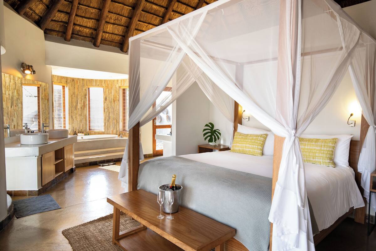 A bedroom with a canopy bed, a bedside table with a champagne bottle, a bathroom with a bathtub, and a large window with blinds.