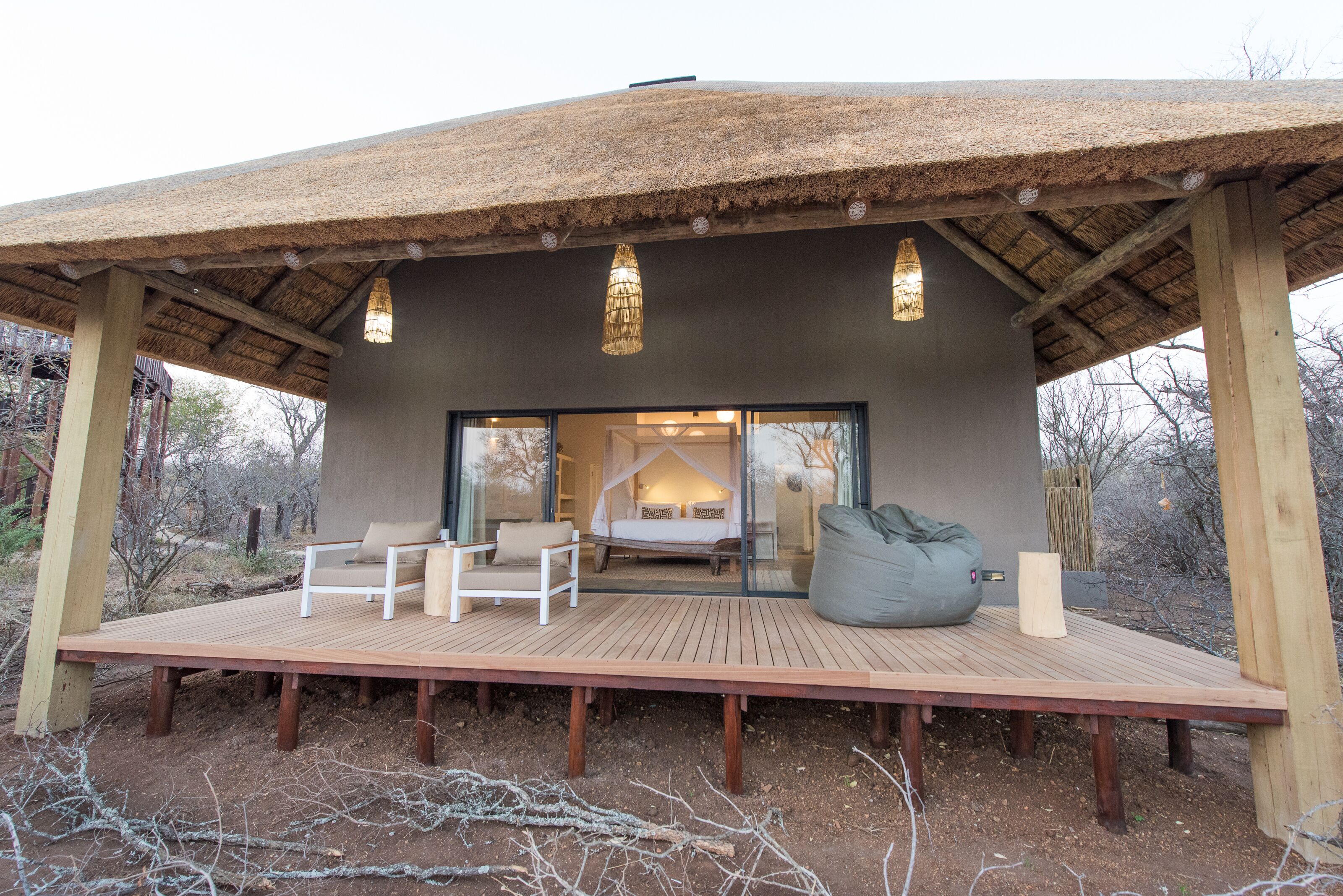 A thatched-roof structure with a wooden deck, a bed with a canopy, and a seating area with chairs and a bean bag.