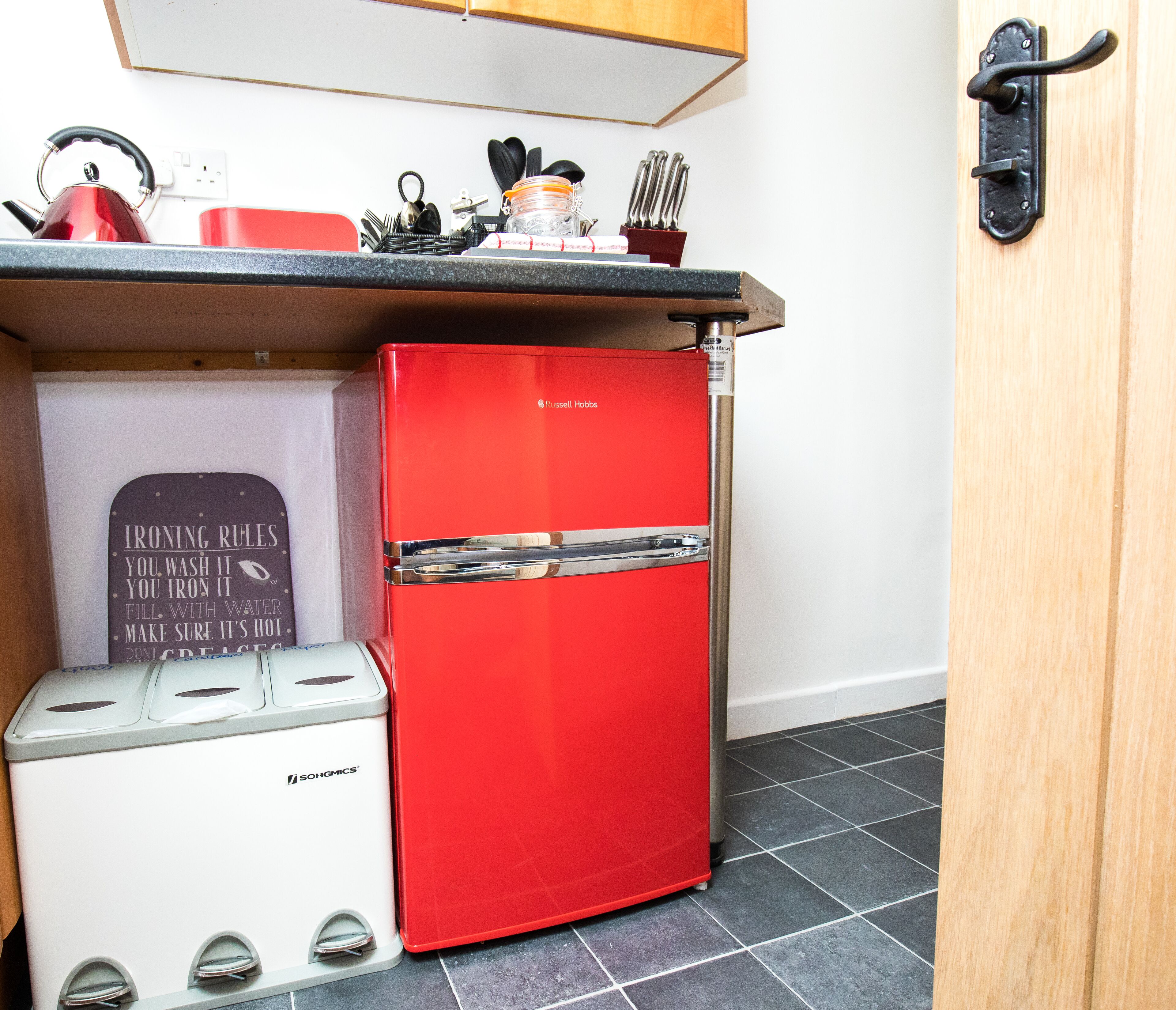 Cocina compacta con un refrigerador rojo, un tostador y un cartel con normas para la plancha.