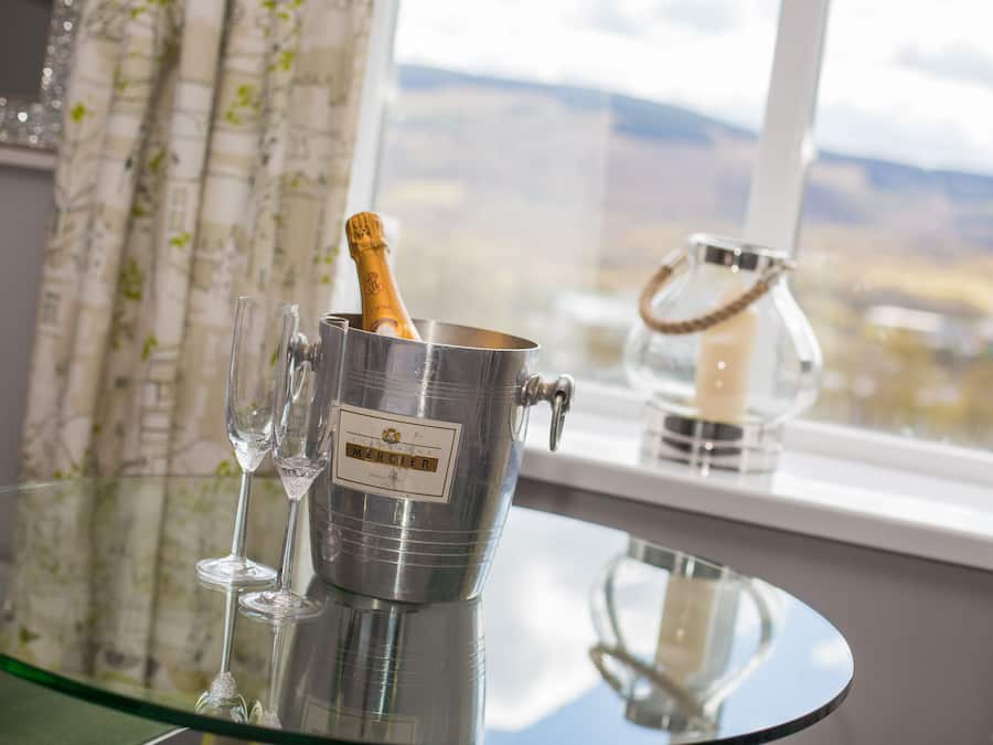 A metallic champagne bucket with a corked bottle on a glass table, near a window with a view of mountains.