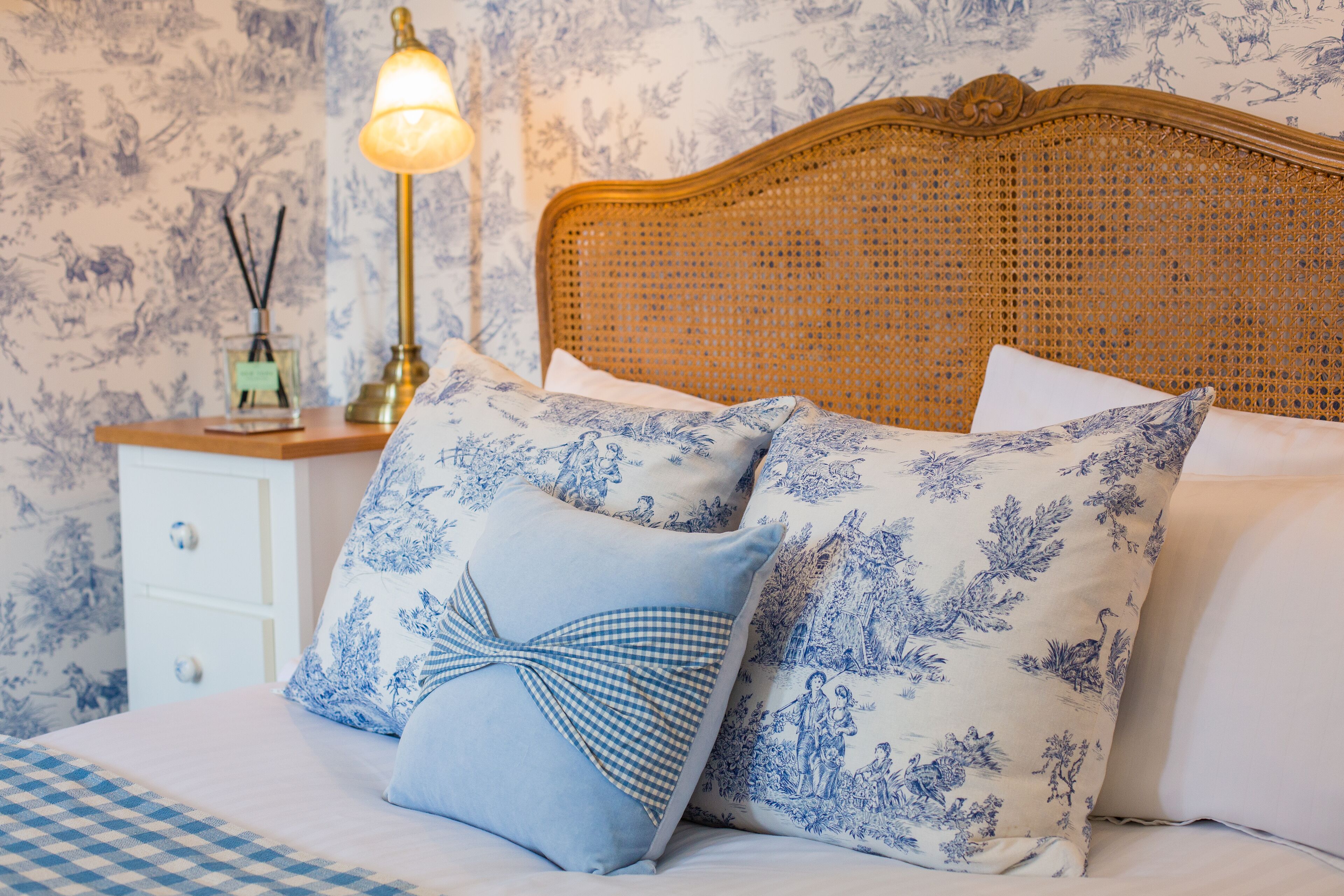 A bedroom with a checkered bedspread, two pillows with a floral pattern, a bedside table with a lamp and a small decorative item.