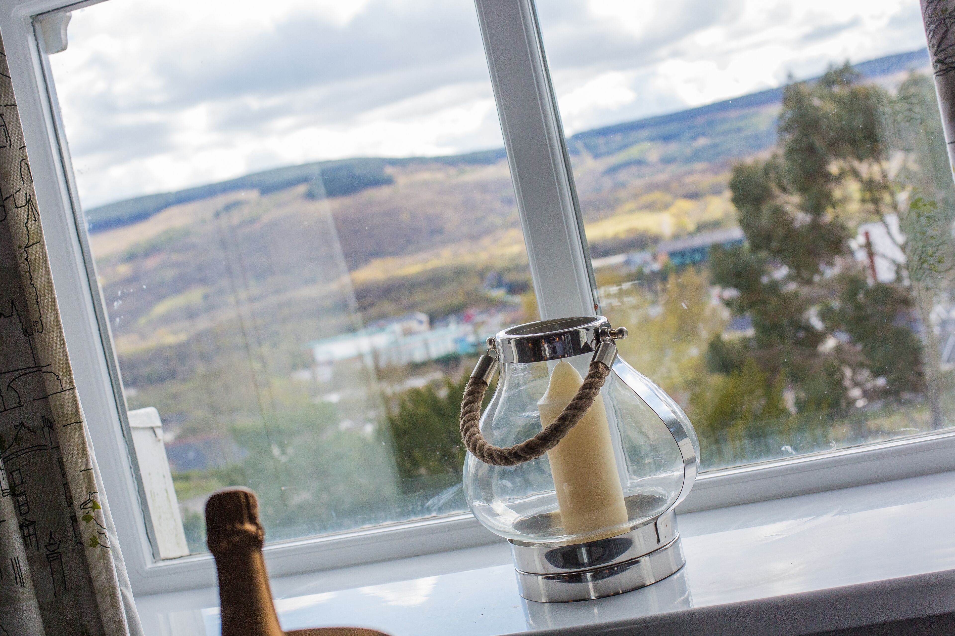 A candle in a glass holder on a windowsill with a view of a landscape.