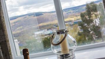 A candle in a glass holder on a windowsill with a view of a landscape.