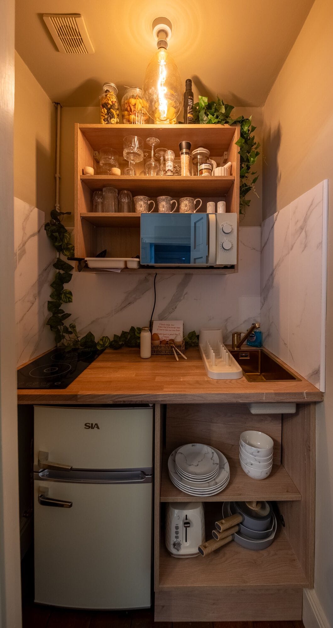 Cocina compacta con microondas, refrigerador y repisas con vasos y frascos.