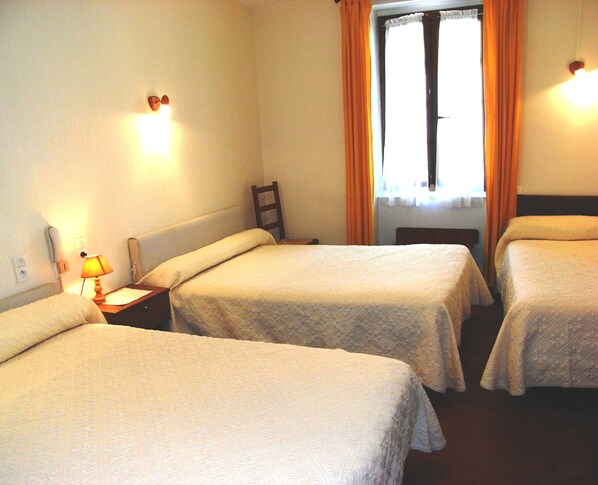 Habitación de hotel con tres camas individuales, una mesita de noche con lámpara, un teléfono y una ventana con cortinas.