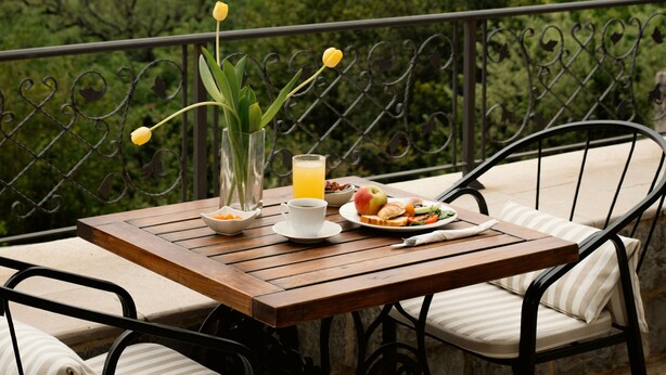 Una mesa de madera con un vaso de jugo de naranja, un bol de fruta y un plato de comida en un balcón con vistas a árboles y una masa de agua.