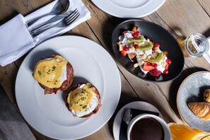 Un desayuno con dos sándwiches huevos benedictinos, una ensalada de frutas, un croissant y una taza de café sobre una mesa de madera.