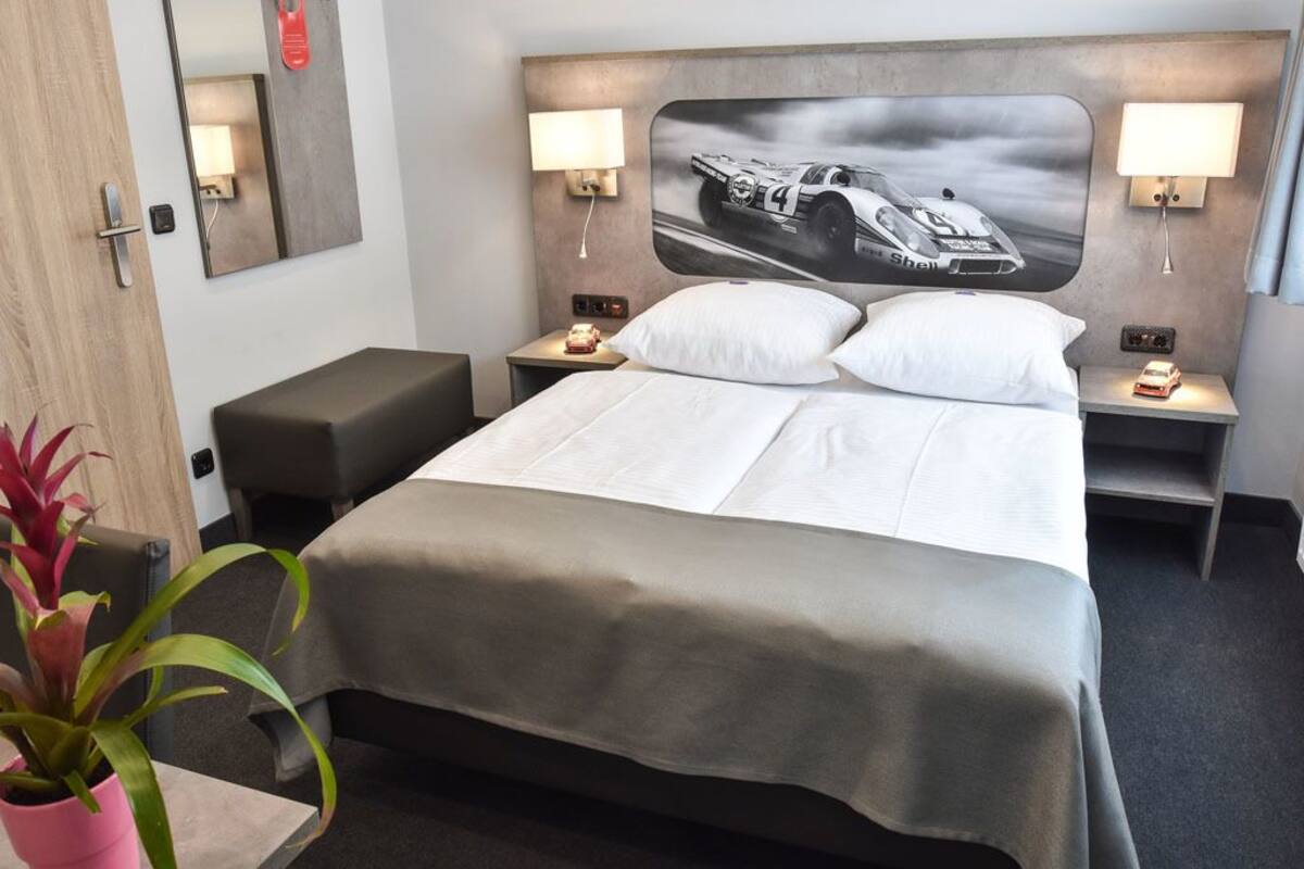 Hotelkamer met een bed, nachtkastjes, lampen, een spiegel, een kamerplant en een wanddecoratie van een auto.