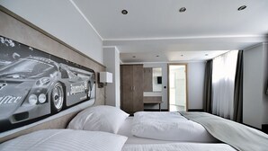 A hotel room with a large bed, a headboard with a car-themed artwork, a mirror, a wardrobe, and a bathroom visible through an open door.