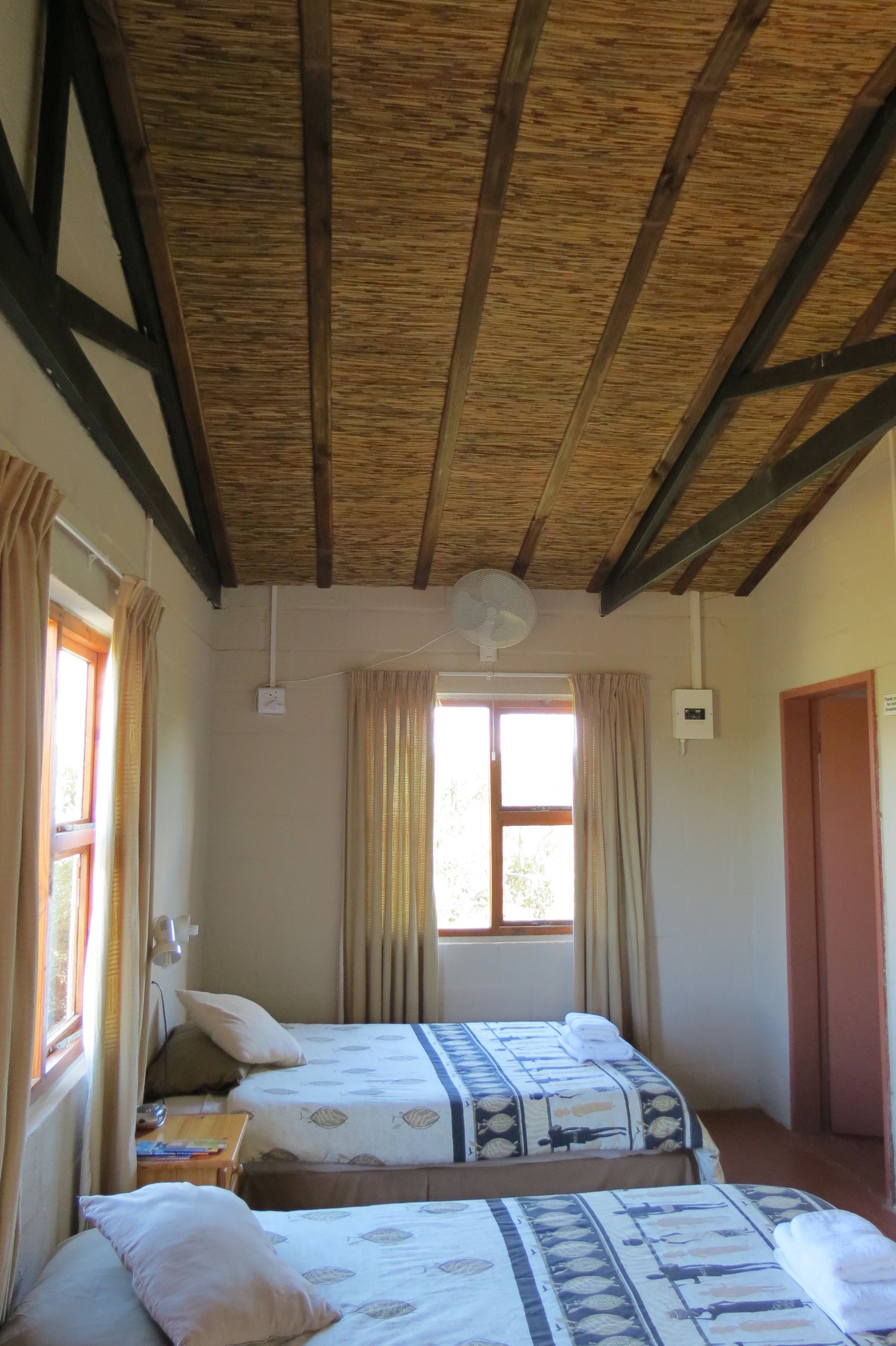 Una habitación con dos camas, vigas de madera en el techo, una ventana con cortinas y un ventilador fijado a la pared.
