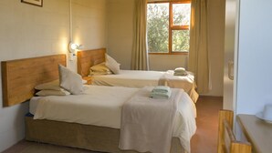 A room with two beds, each with a wooden headboard, a bedside table, and a lamp. There is a window with curtains, and a small table with towels on it.