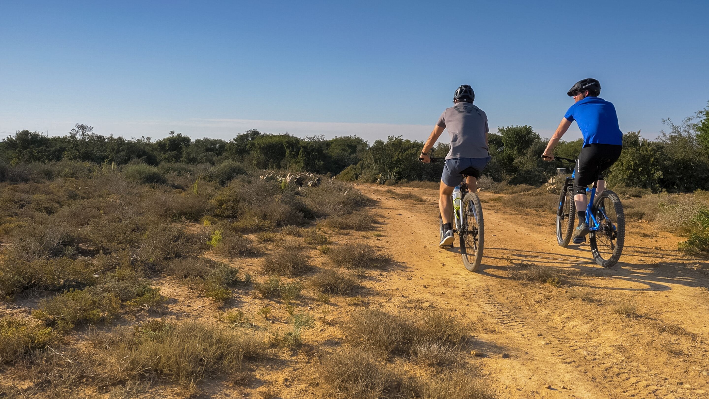 Deux personnes à vélo sur un chemin de terre, entourées d’une végétation sèche et d’arbres.