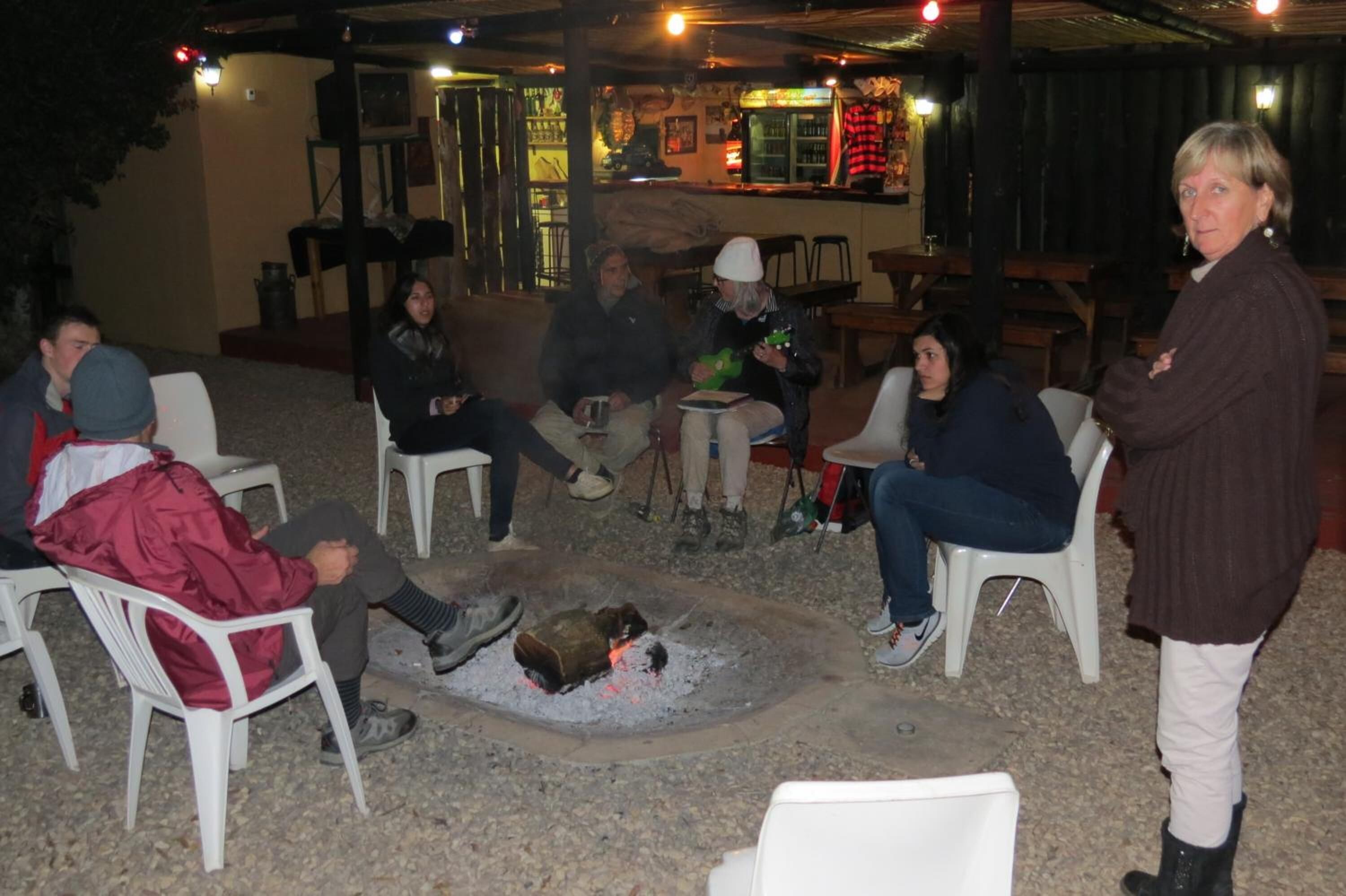 Un groupe de personnes est assis autour d’un foyer dans un espace extérieur aménagé avec des tables et des chaises.