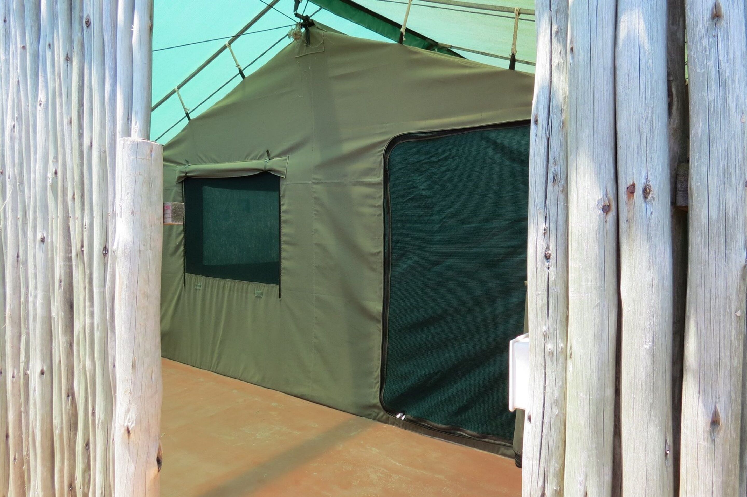 Tienda de campaña con interior verde y una ventana, montada frente a una estructura de madera.