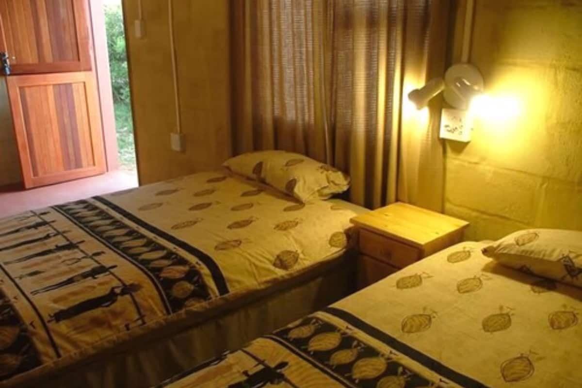A room with two beds, each with a leaf-patterned bedspread, a wooden nightstand, and a wall-mounted light fixture.