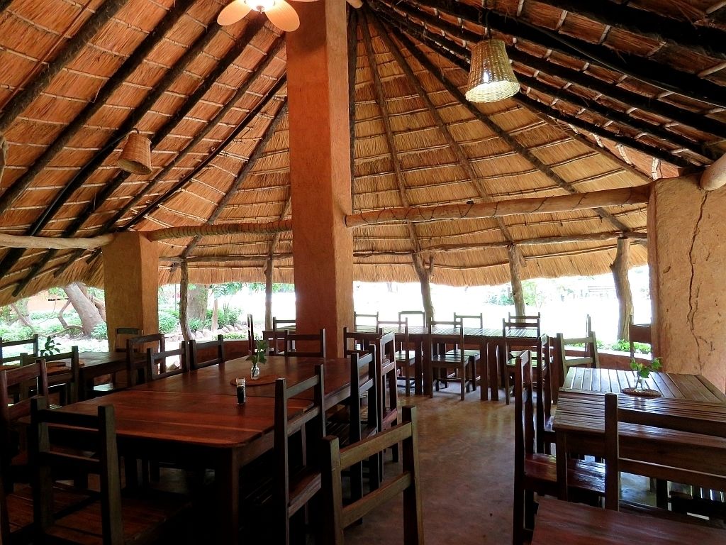 An indoor dining area with thatched roof ceilings, wooden tables and chairs, and large windows allowing natural light.