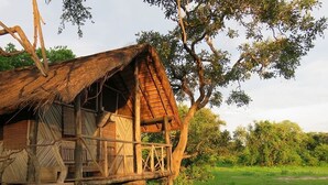 A rustic wooden structure with a thatched roof, elevated on stilts, surrounded by lush greenery. - Croc Valley Camp (South Luangwa National Park)