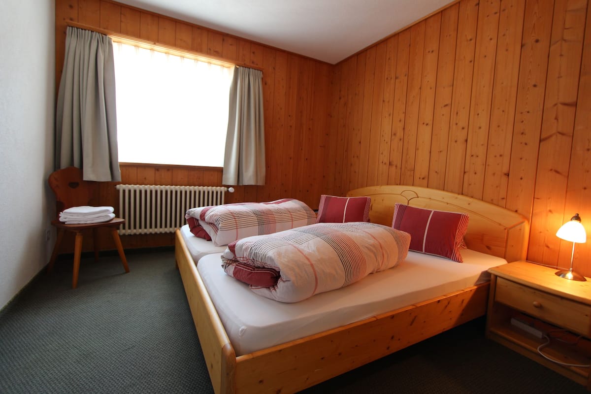 A wooden-paneled room with a bed, a chair, a nightstand, and a window with curtains.