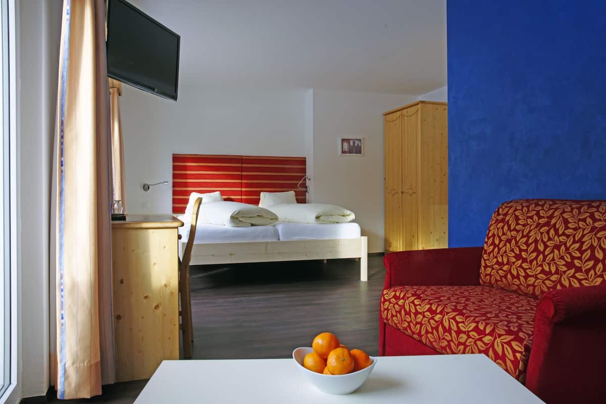 A hotel room with a bed, a red sofa, a TV, and a bowl of oranges on a table.