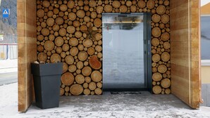 A wooden cabin with a door and a small black trash bin outside. - Hotel Al Rom (Val Muestair)