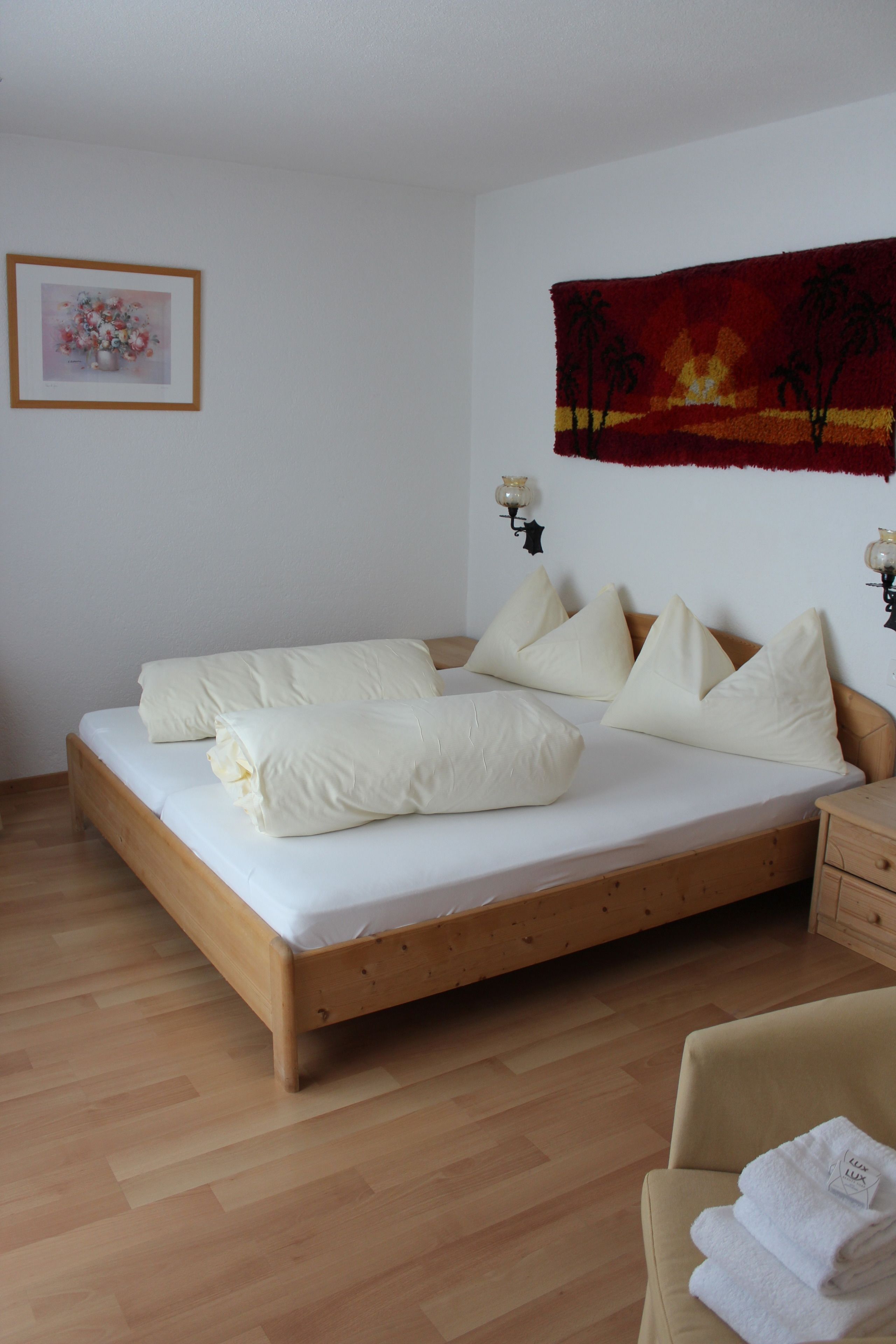 Un letto rifatto con lenzuola e cuscini bianchi, una testiera in legno, un comodino con una lampada e una sedia con asciugamani piegati.