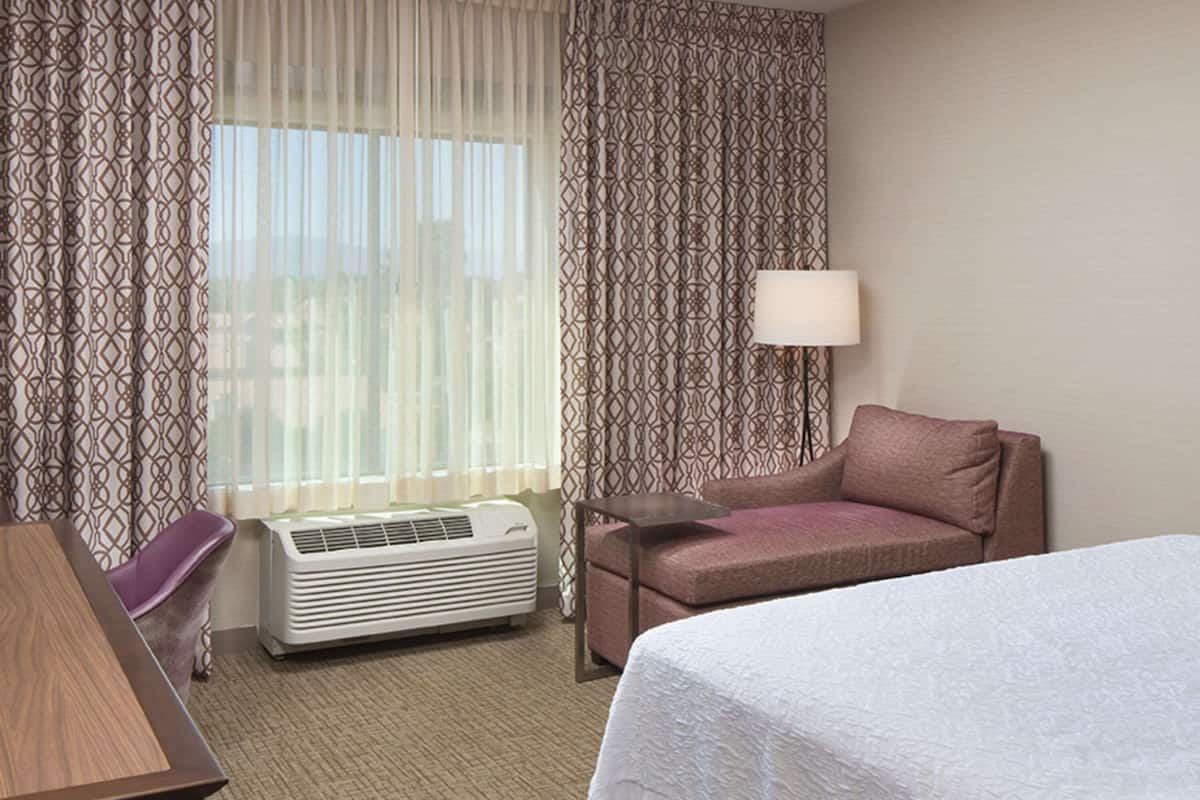 A hotel room with a bed, a desk, a chair, a sofa, a lamp, and an air conditioner.