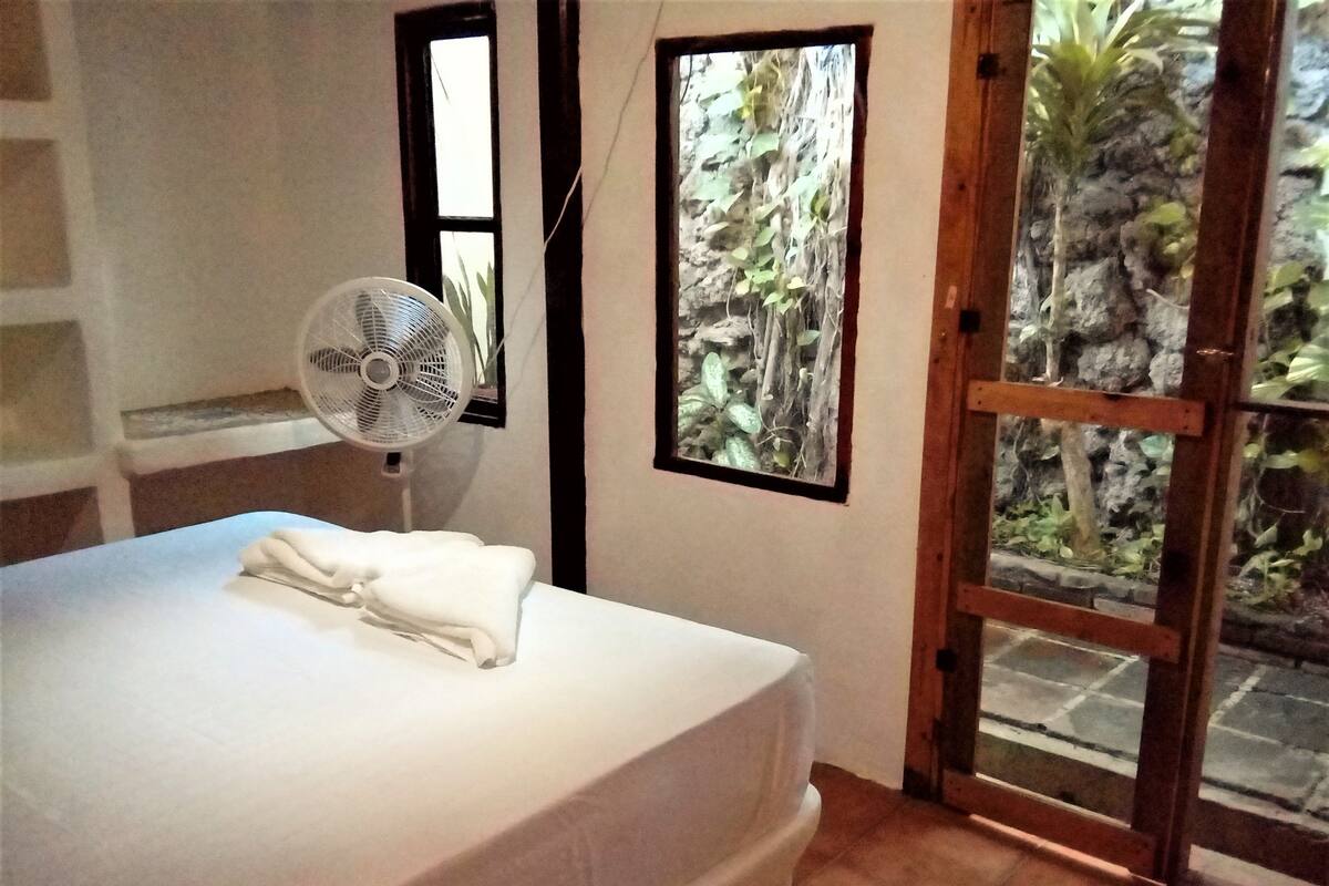 A bedroom with a bed, a fan, and a view of a garden through a glass door.