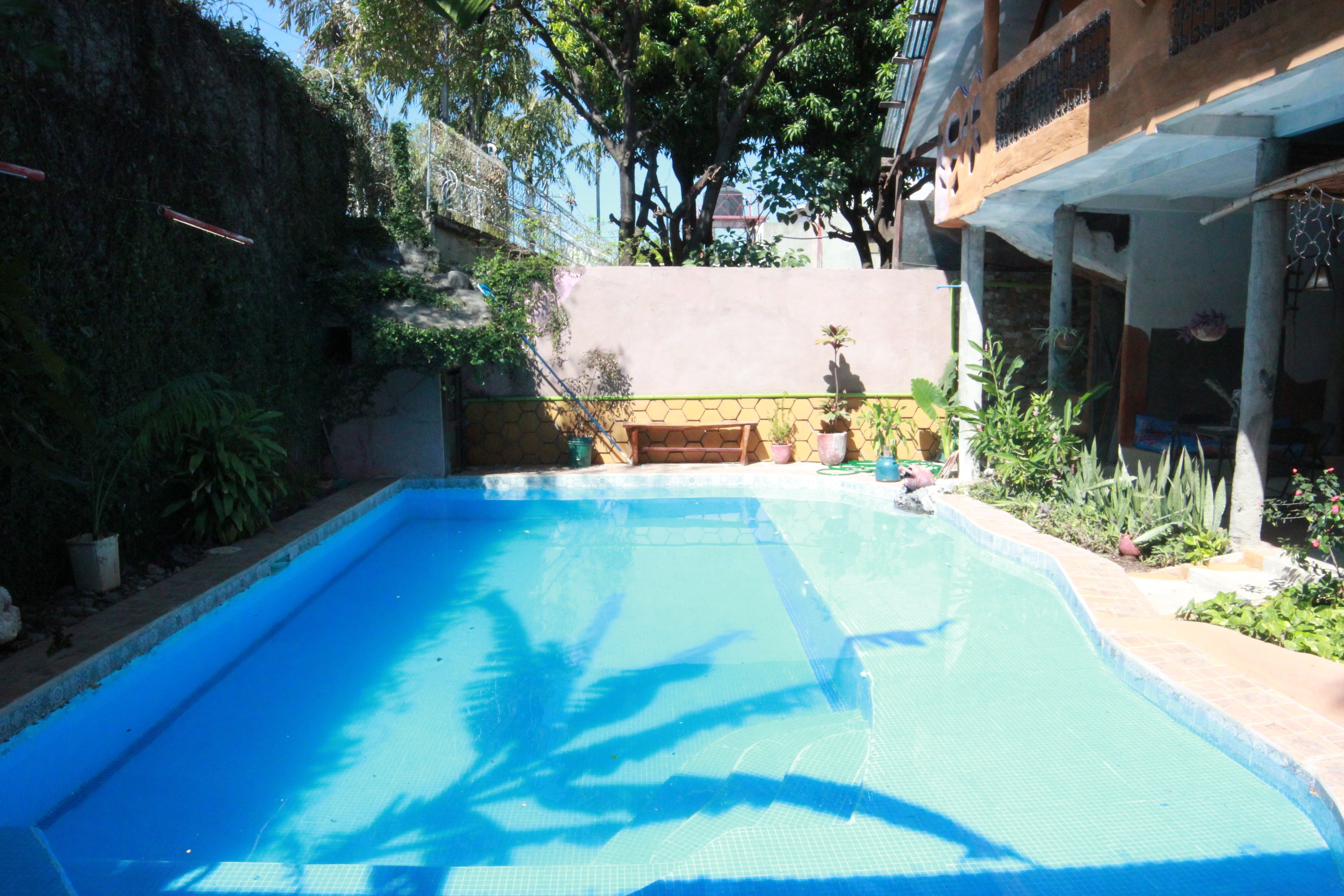 A swimming pool with a tiled edge, surrounded by potted plants and a building with a balcony.