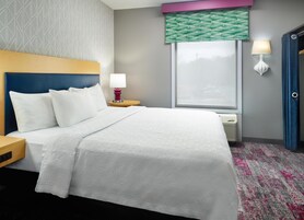 Habitación de hotel con cama, mesita de noche, lámpara, ventana con vistas y puerta.