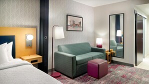 Ein Hotelzimmer mit einem Bett, einem grünen Sofa, einem kleinen Tisch mit einer Lampe, einem Spiegel und einem Stadtbild an der Wand.