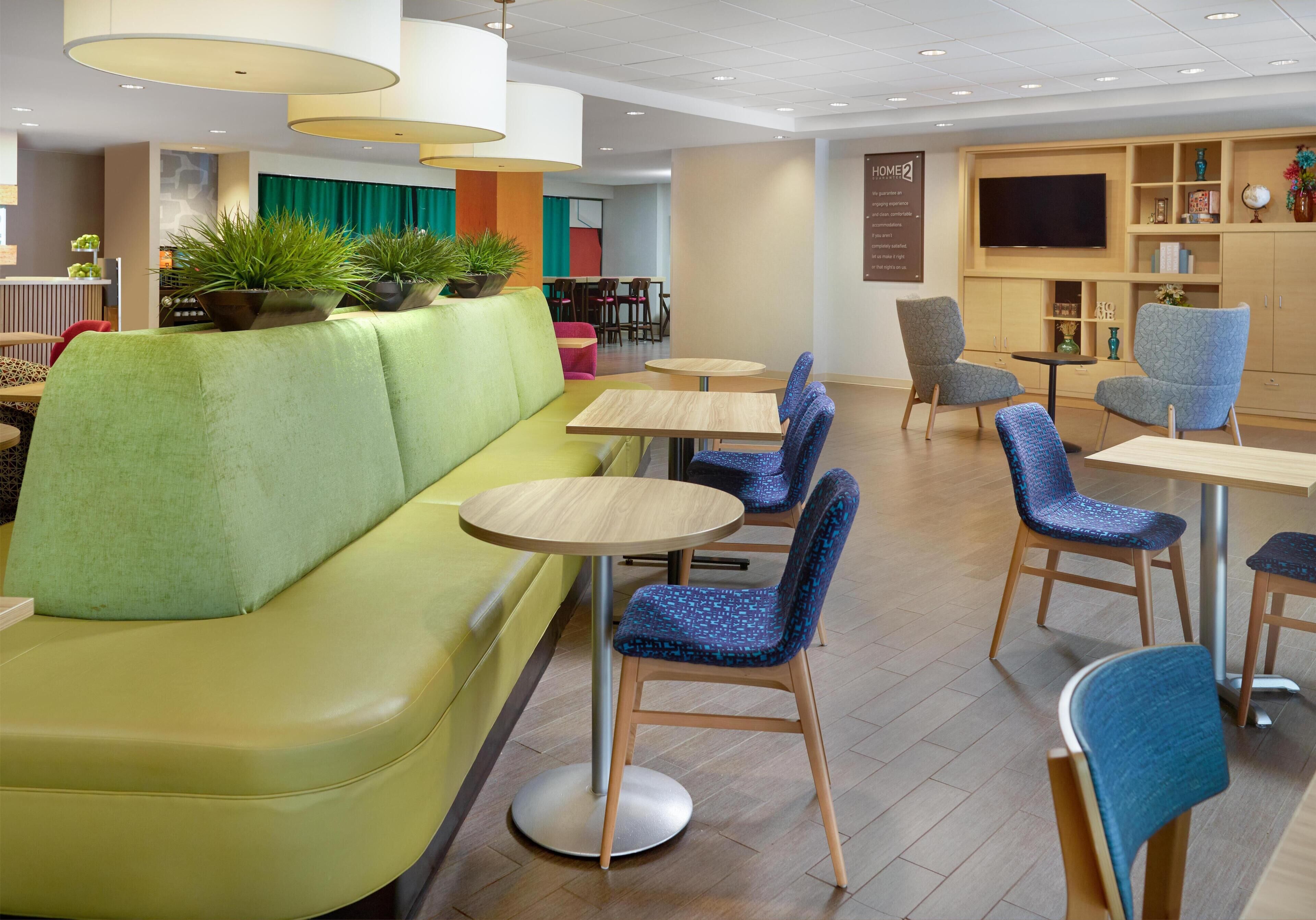 Un hall d’hôtel moderne avec un canapé vert, des tables rondes, des chaises bleues et un téléviseur fixé au mur.