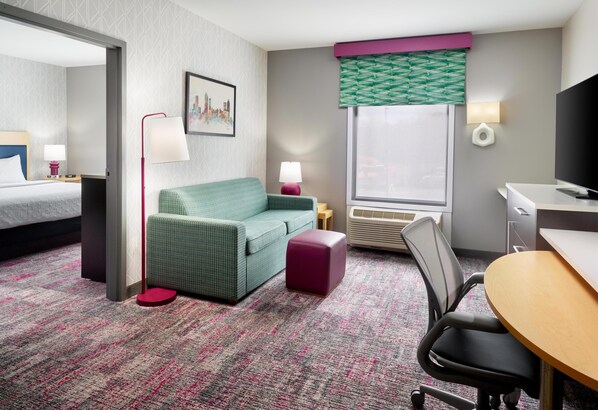 A hotel room with a bed, a green sofa, a small table, a desk, a chair, a TV, a window with blinds, a wall-mounted lamp, and a picture on the wall.