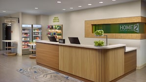 Ein Hotel-Empfangsbereich mit einem Holzbüro, einem 'Home 2'-Schild und einem Snack-Kühlschrank.