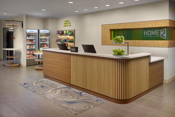 A hotel reception area with a wooden reception desk, a 'Home 2' sign, and a snack refrigerator. - Home2 Suites Newnan (Newnan)