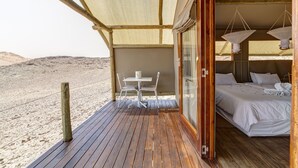 A wooden deck with a table and chairs, a bed with white linens, and a view of the desert. - Kanaan N/a'an ku sê Desert Retreat (Kanaan)