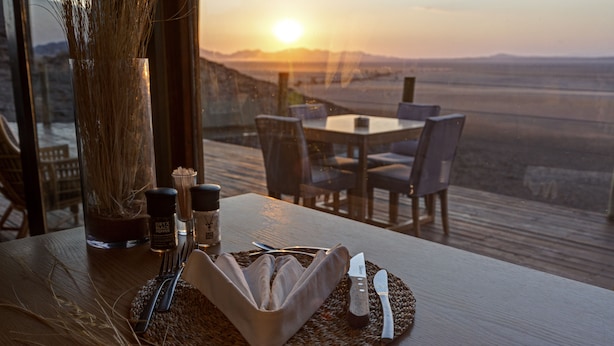 A dining table with a view of the sunset, a vase with dried plants, and a bottle of Oryx Black Gin. - Kanaan N/a'an ku sê Desert Retreat (Kanaan)