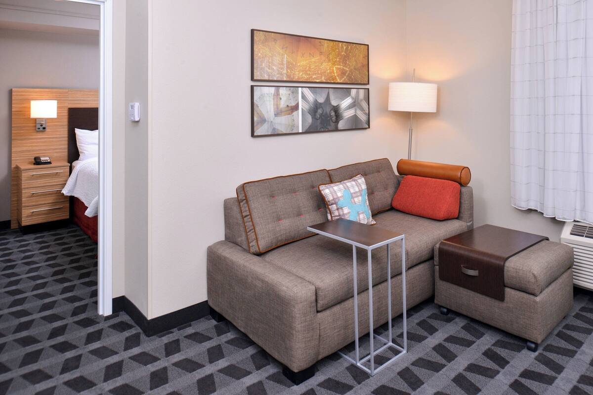 A hotel room with a sofa, a small table, a lamp, and a wall-mounted artwork.