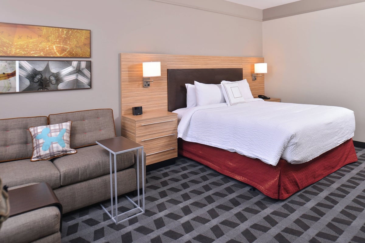A hotel room with a bed, a nightstand, a bedside table, a sofa, and a wall-mounted artwork.