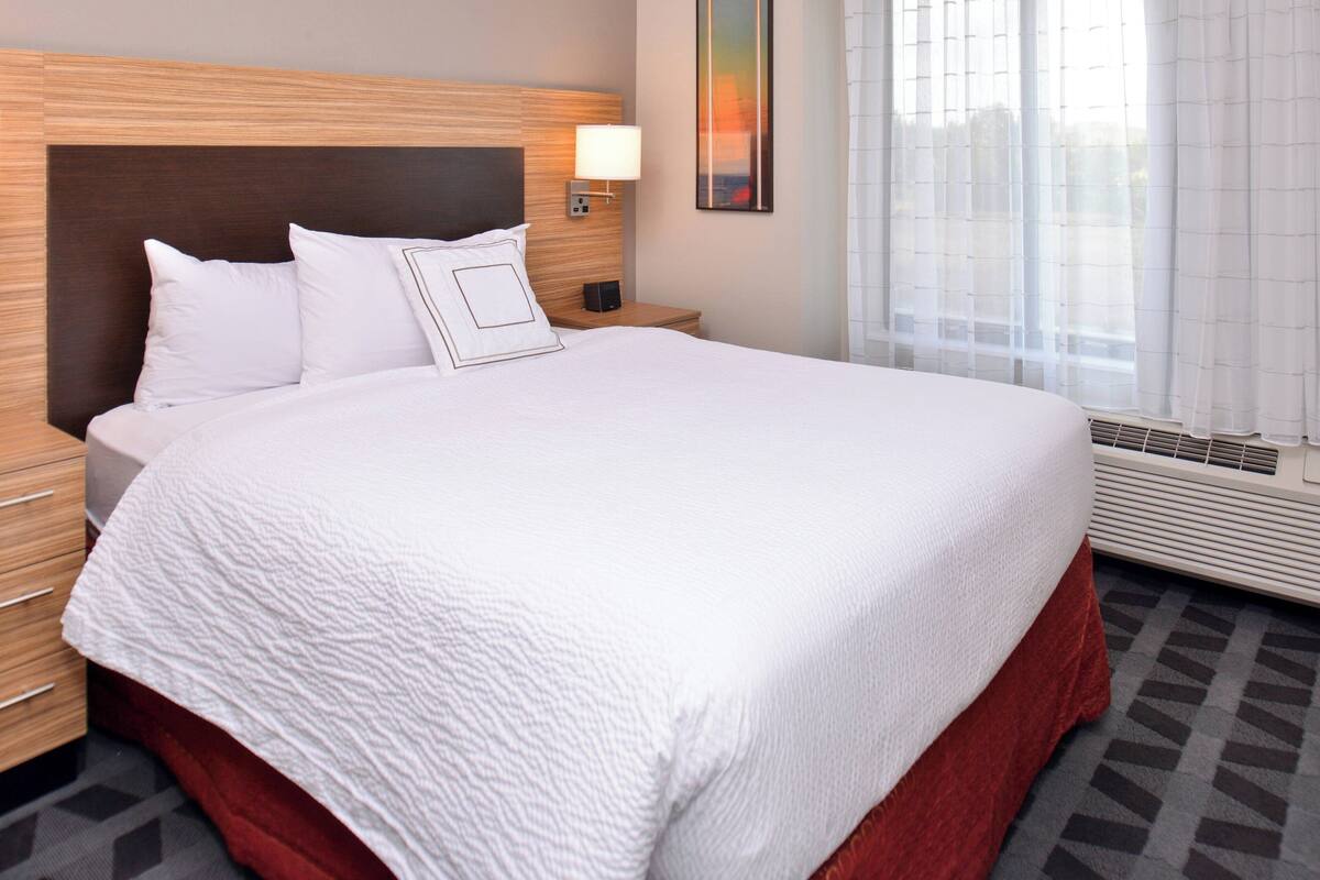 A hotel room with a large bed, a wooden headboard, a bedside table with a lamp, and a window with sheer curtains.