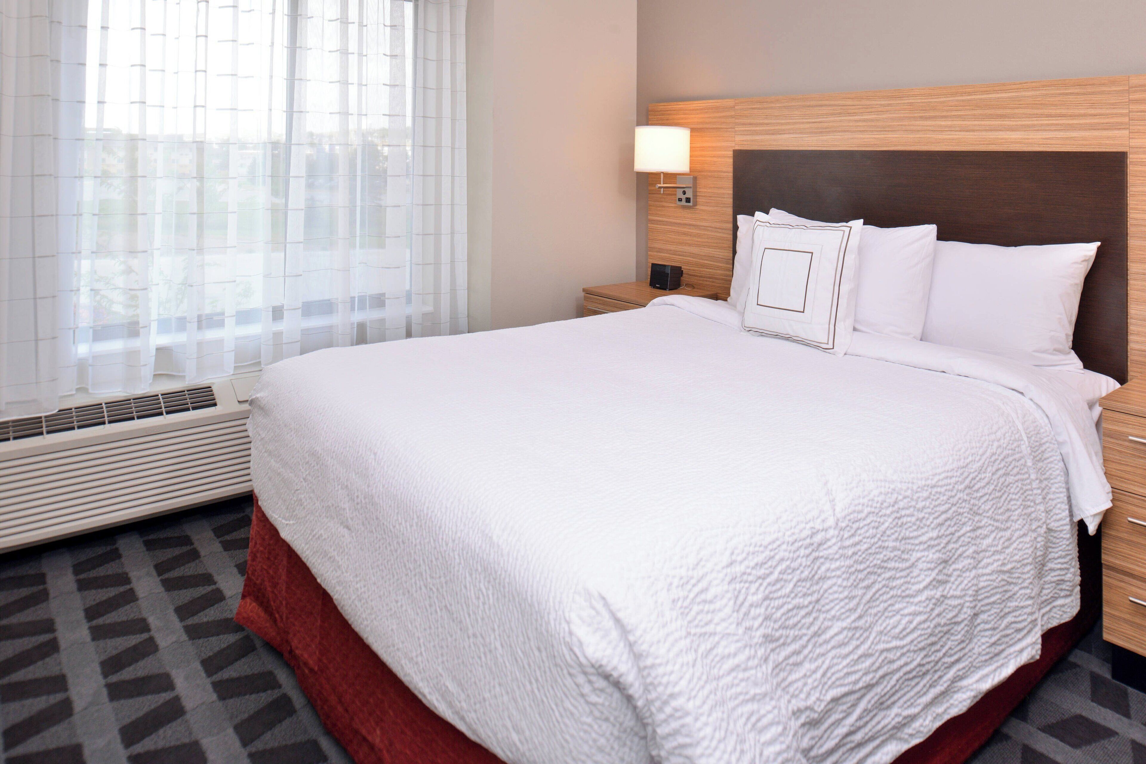 A hotel room with a large bed, a bedside table, a nightstand, a wall-mounted lamp, and a window with sheer curtains.